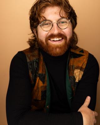 Jacob smiles warmly, wearing round glasses, a black turtleneck, and a patterned vest against a warm tan background.