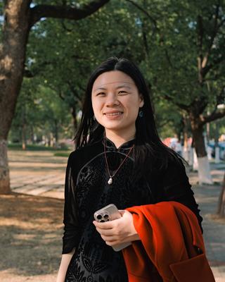 Xiaorui Zhou smiling outdoors beneath leafy trees, wearing a black patterned outfit and holding a phone and orange coat.