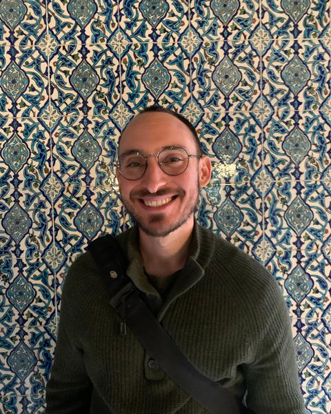 Mitchell Bacci smiles warmly before intricate blue patterned tiles, wearing round glasses, green sweater, and crossbody strap.