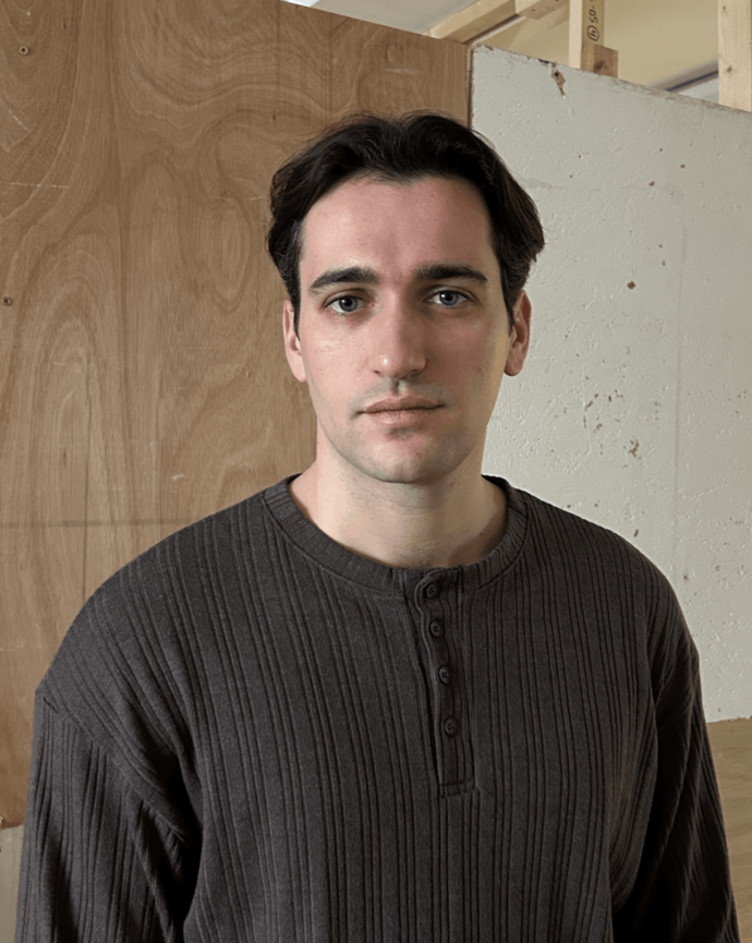 Francesco with dark hair wearing ribbed brown shirt stands indoors against unfinished wall and wooden panel background.