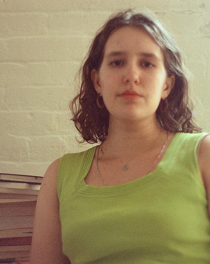 Ida Beckett wearing a lime green sleeveless top, seated indoors beside stacked books against a softly textured stone wall.